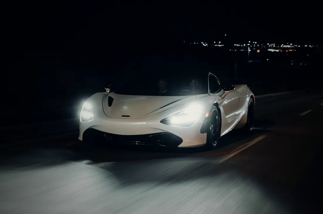 A white McLaren 720S driving towards the camera at night, headlights on