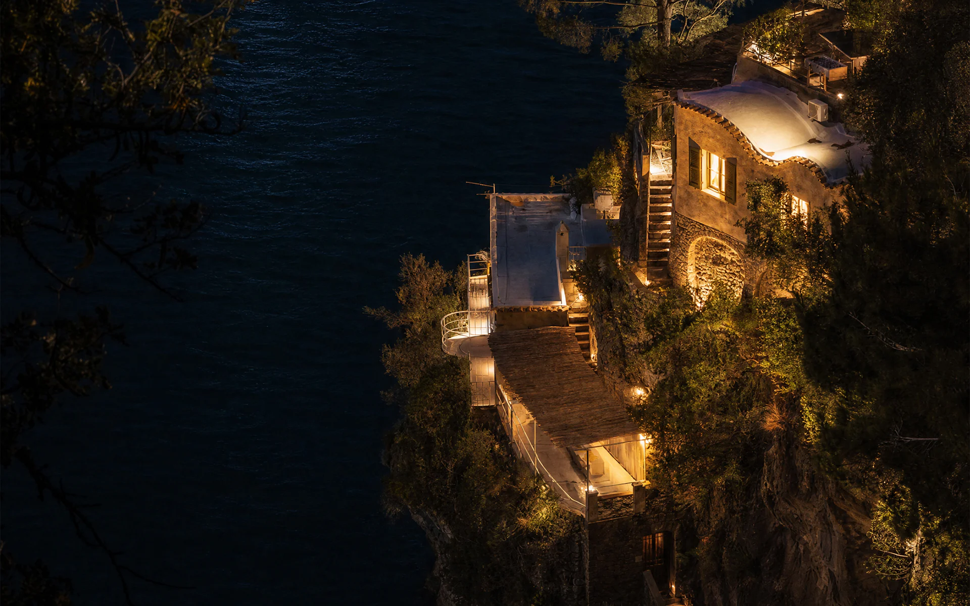 Stone villa with lit terraces stepping down to a dark sea at night